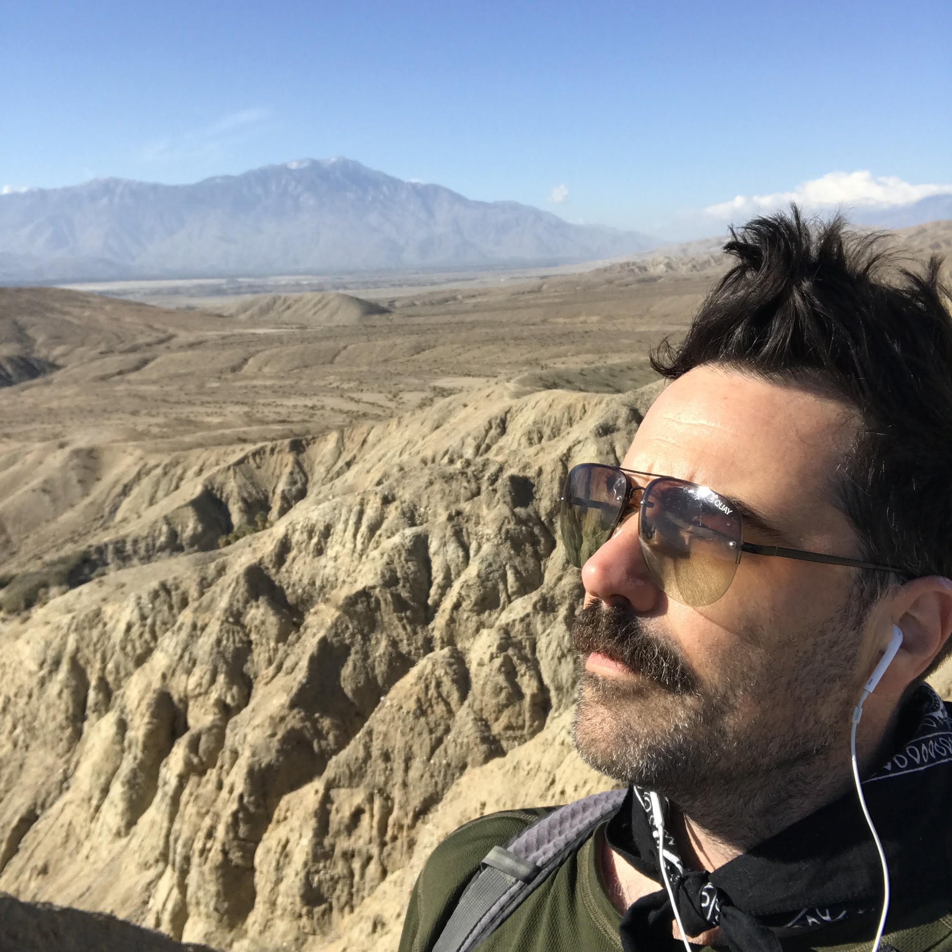Man wearing sunglasses and earbuds looking out over a desert landscape with mountains
