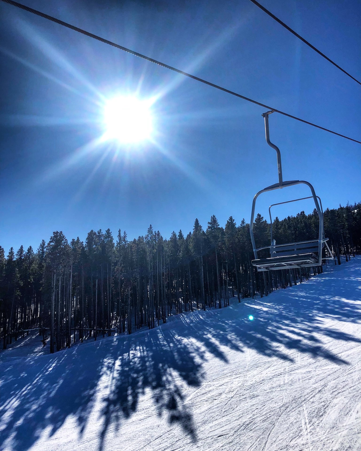 A ski lift chair on a cable above a snowy slope with evergreen trees and a bright sun.