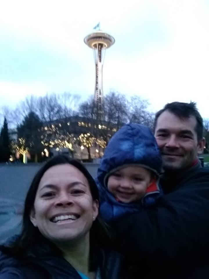 Selfie of a woman, child, and man in winter coats in front of the Seattle Space Needle at dusk