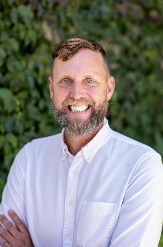 A smiling bearded man in a white shirt with arms crossed, standing in front of green foliage.