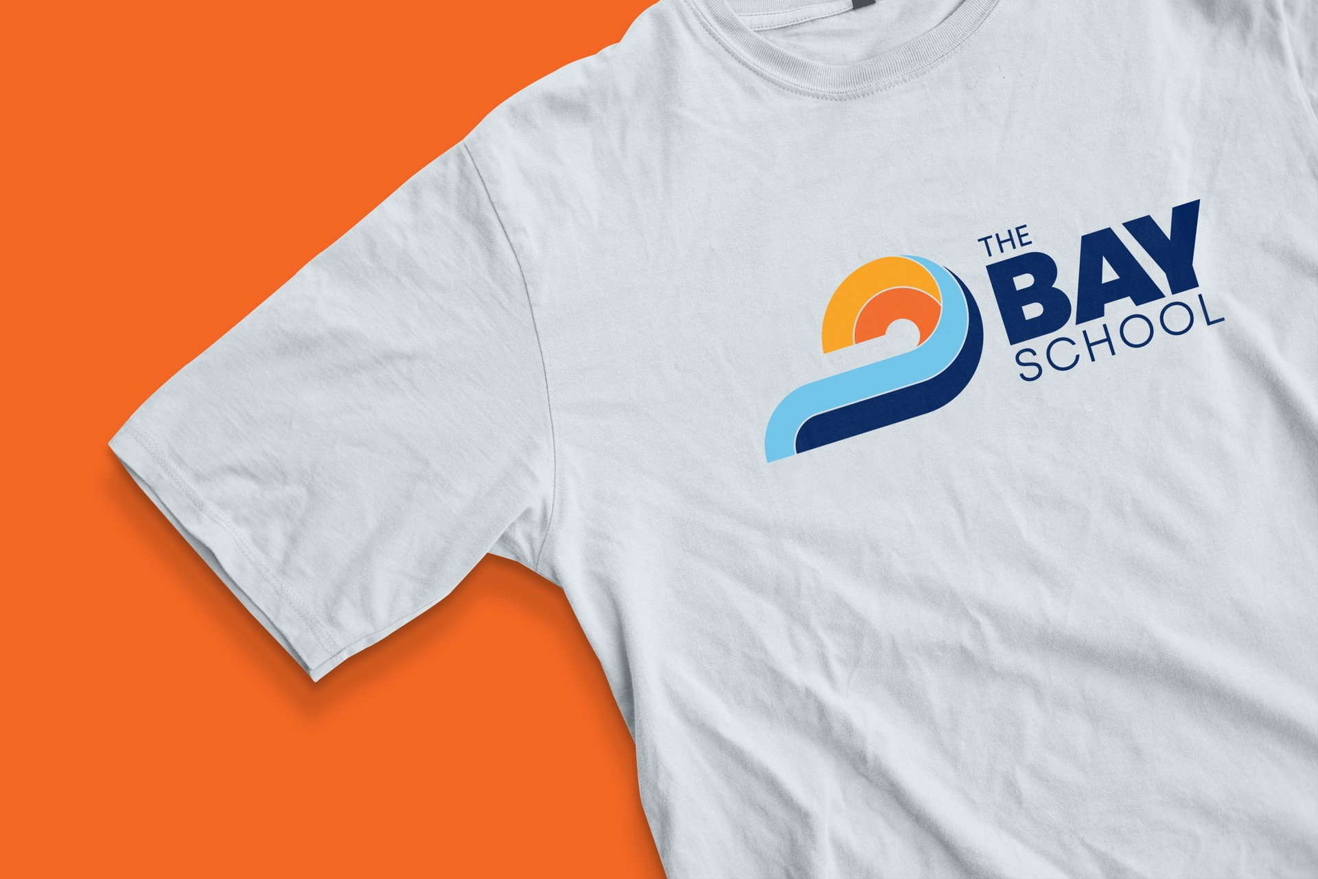 Light blue t-shirt printed with The Bay School wave-and-sun logo on an orange background
